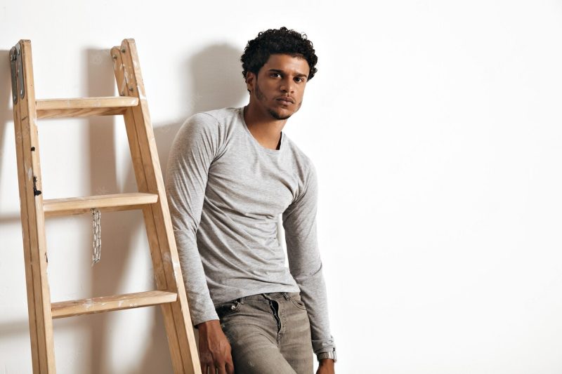 Serious sad muscular African American model in heather grey long-sleeve cotton t-shirt and jeans leaning on white wall next to a wooden stepladder Free Photo