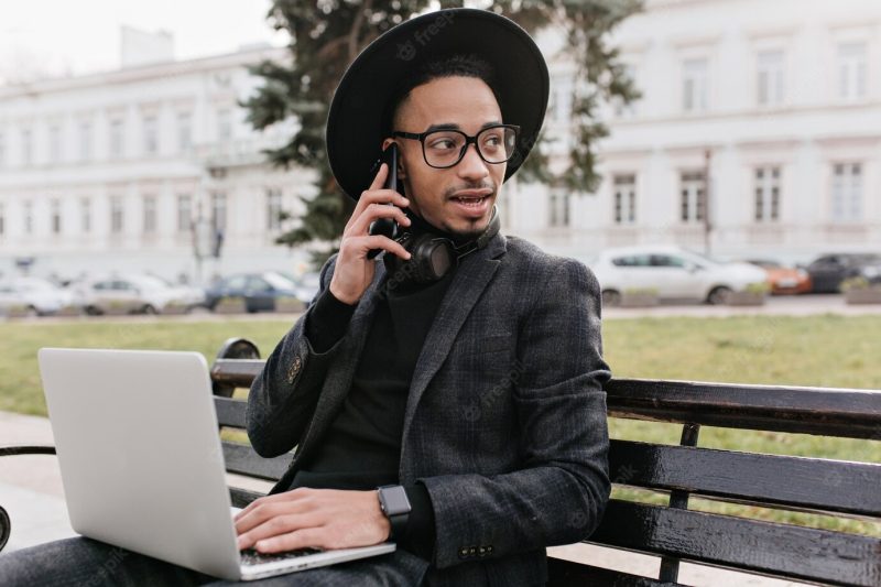 Serious young businessman talking on phone while working with computer in park. outdoor photo of busy african guy using laptop in square. Free Photo