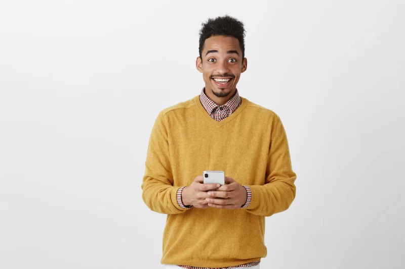 Sharing great news with friend. portrait of happy thrilled dark-skinned man in trendy outfit, holding smartphone, gazing with broad smile, being on cloud nine from happiness Free Photo