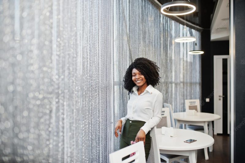 Cheerful business African American lady with afro hair wear white blouse and green pants posed in cafe Free Photo