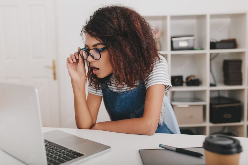 Shocked young woman in striped shirt looking at laptop screen and holding glasses Free Photo