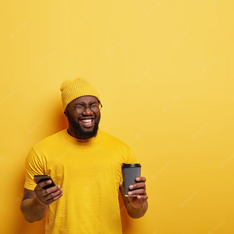 Shot of happy pleased dark skinned guy closes eyes from pleasure, wears yellow stylish hat and t shirt, messages in social networks, holds modern cellular and coffee to go, giggles at something funny Free Photo