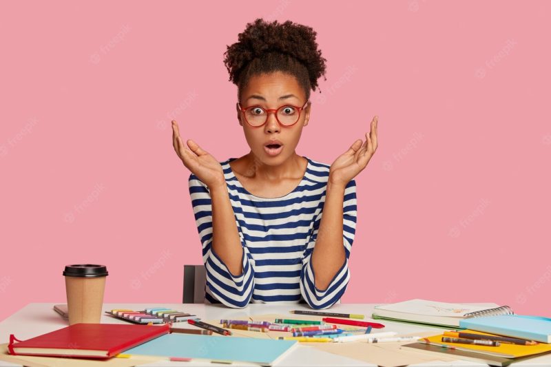 Shot of impressed terrified woman with curly hair, keeps hands near face, looks in terror, poses at workplace, has puzzled expression, works as illustrator, drinks hot beverage, models indoor Free Photo