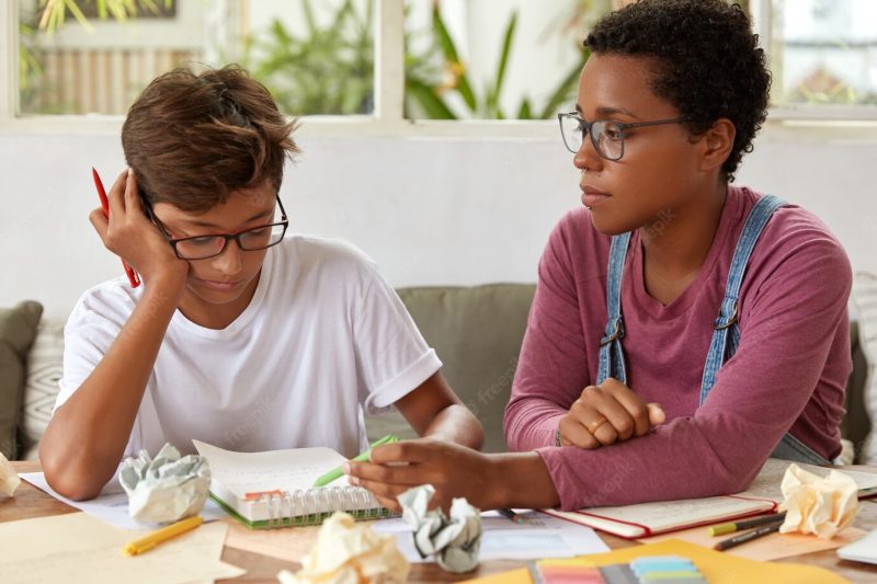 Shot of mixed race guy and female sit together at workplace, discuss ideas for project, wear glasses. black woman with piercing explains something to brother, indicates at writings in diary. Free Photo