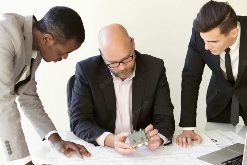 Shot of working process at construction design bureau. three men in office clothes examining new perspective project. main design engineer sitting at table, his coworkers standing aside of him. Free Photo