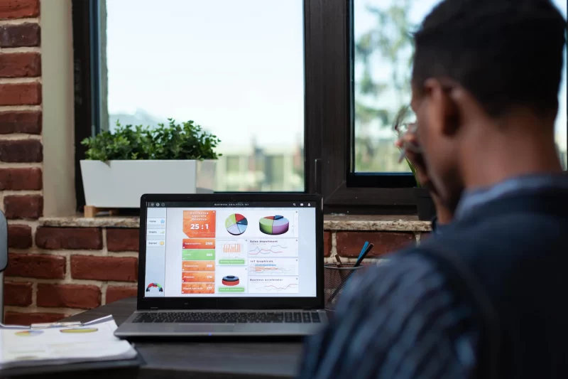 Over shoulder view of african american startup employee looking at laptop screen with business analytics charts sitting at desk. close focus on portable computer display with sales results. Free Photo