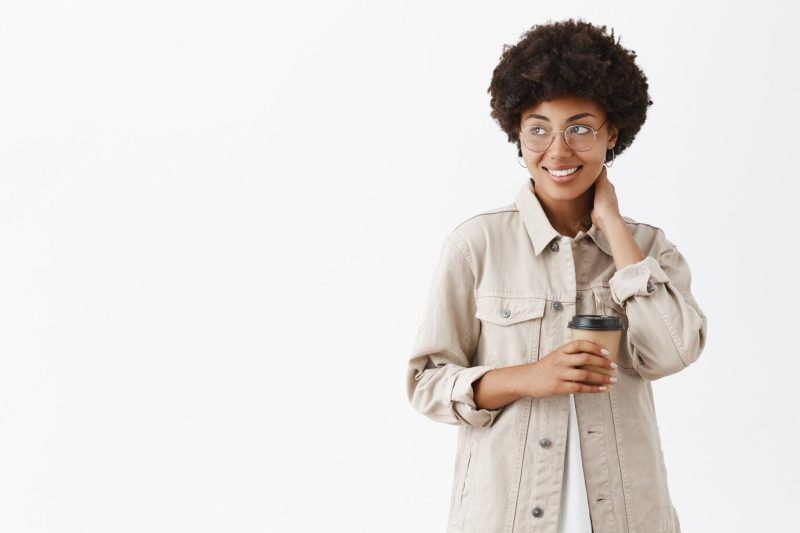 Shy African American female office worker in shirt and glasses touching neck, gazing left with smile and holding paper cup Free Photo