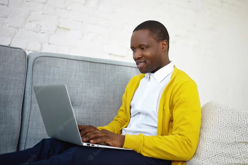 Sideways shot of attractive talented dark skinned male copywriter in stylish wear sitting on couch at home with portable computer on his lap, keyboarding new article for online internet magazine Free Photo