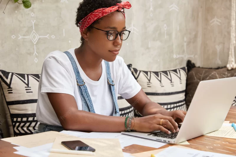 Sideways shot of dark skinned female worker has business online, keyboards on laptop computer Free Photo