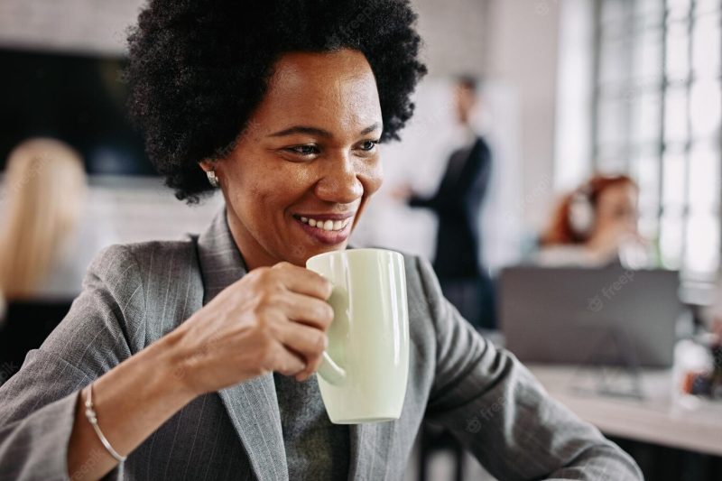 Smiling African American businesswoman drinking coffee while being at work there are people in the background Free Photo