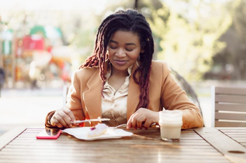 Smiling african american woman drinking coffee and eating dessert in outdoor cafe Free Photo