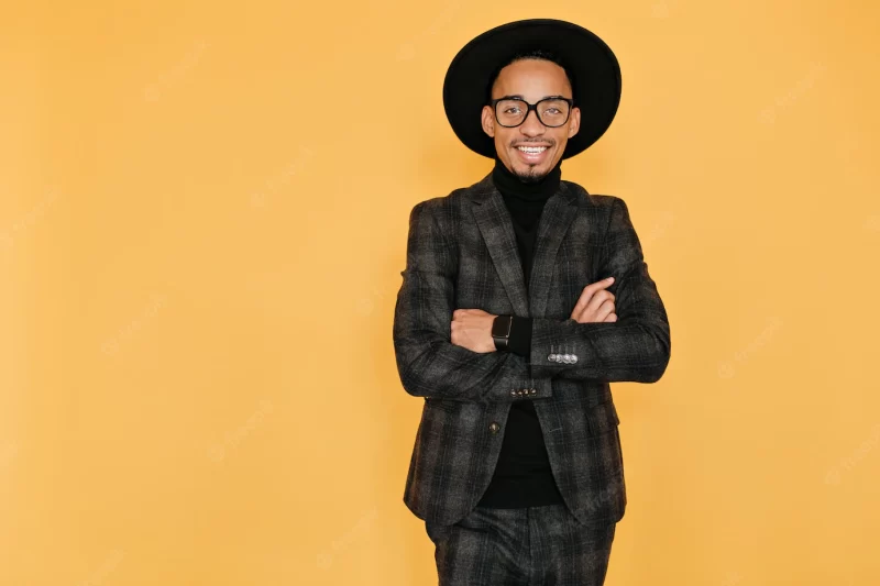 Smiling african guy with inspired face expression. indoor photo of pleased black man in hat standing with arms crossed on yellow wall. Free Photo