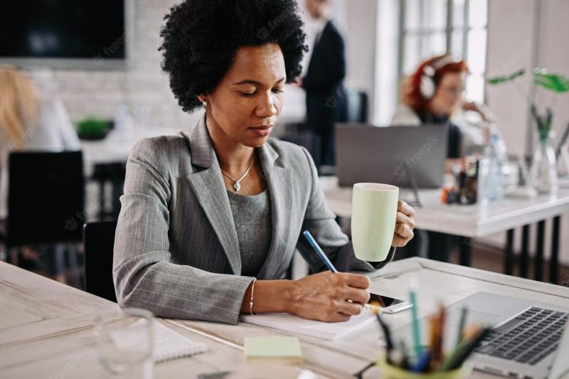 Smiling black business woman with cup of coffee working at her desk and taking notes in her notebook there are people in the background Free Photo