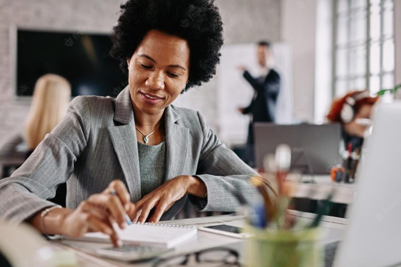 Smiling black entrepreneur working on financial reports at her desk in the office there are people in the background Free Photo