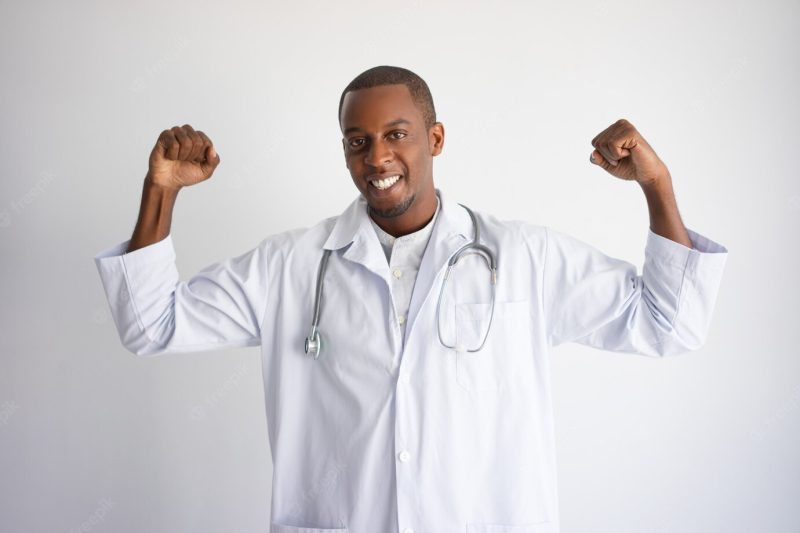 Smiling black male doctor pumping fists and celebrating success. Free Photo