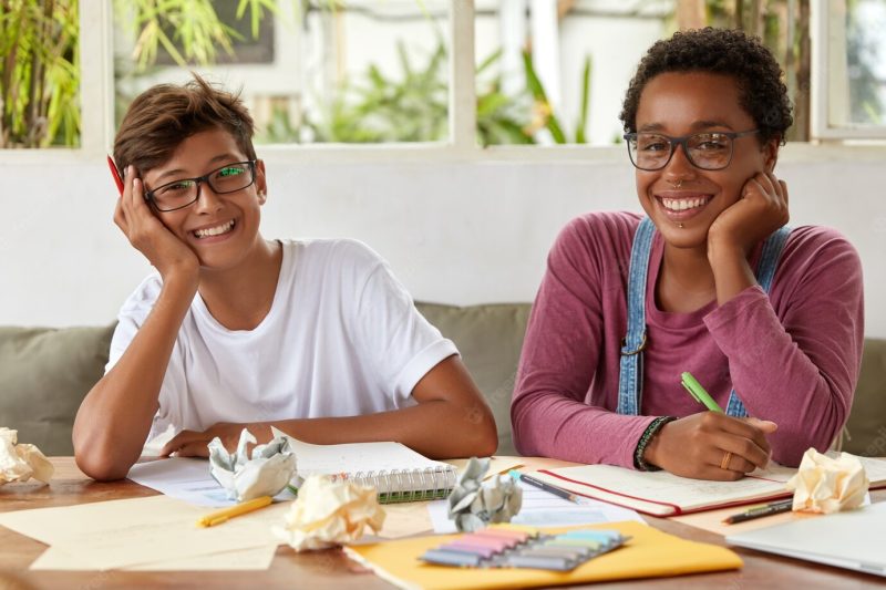 Smiling dark skinned woman gives good advice to male classmate, talk about common homework, write down records in spiral notebook, talk about common project and make research or plans together Free Photo