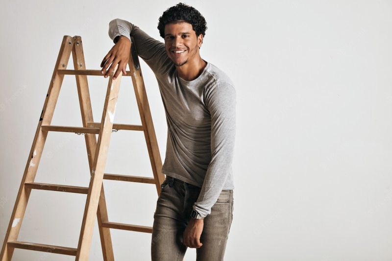 Smiling relaxed tall muscular young model wearing a plain heather gray longsleeve t-shirt and slim gray jeans leaning on wooden stepladder isolated on white. Free Photo