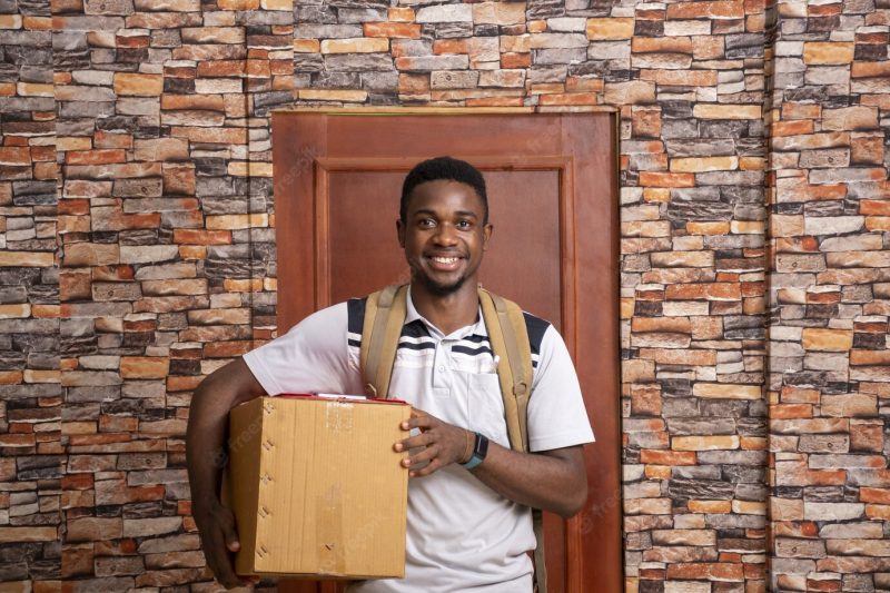 Smiling young african courier holding a package standing in front of a door Free Photo