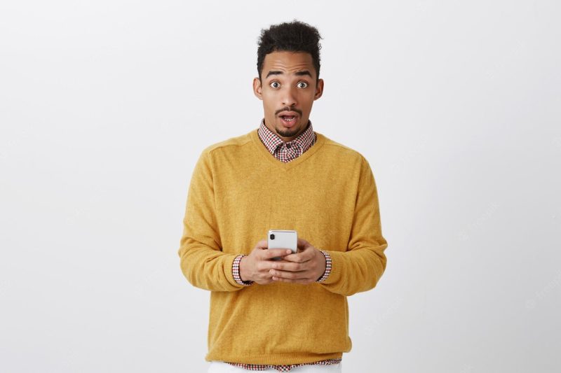 Someone tried to hack his phone. shocked handsome man with afro hairstyle in trendy clothes holding smartphone, staring with amazed expression, being surprised over gray wall Free Photo