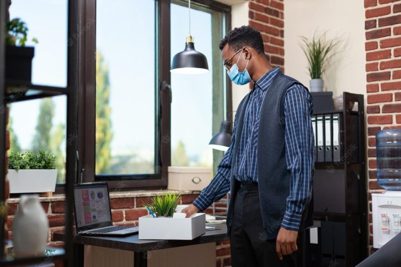 Startup employee wearing covid mask putting personal belongings in tray giving a last look at his desk with laptop. laid off office worker looking with regret at portable computer. Free Photo