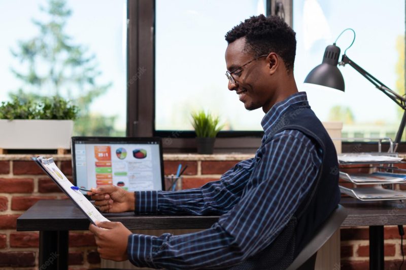 Startup owner holding clipboard with annual bussiness charts happy with turnover results sitting at desk with laptop. entrepreneur comparing key performance indicators on portable computer. Free Photo