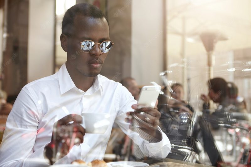 Staying connected. view through window glass of handsome black european employee in shades and formal shirt drinking coffee during lunch break and checking e-mail on touchscreen mobile phone Free Photo