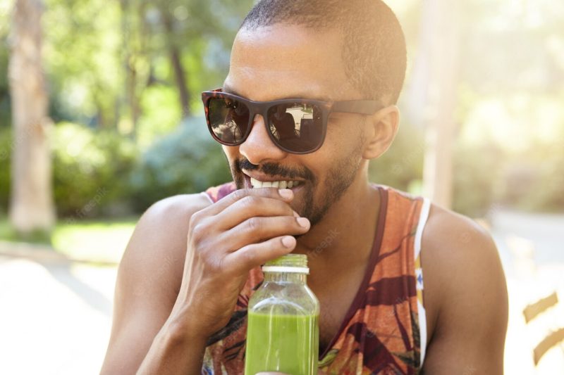 Street lifestyle concept. young smiling African American male with mustache and short beard drinking fresh juice during date, dressed casually in colorful tank top and trendy shades or sunglasses Free Photo