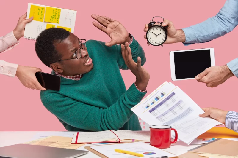 Stressful dissatisfied dark skinned guy makes refusal sign, raises palms over alarm clock, touchpad and papers, has many tasks in one minute Free Photo