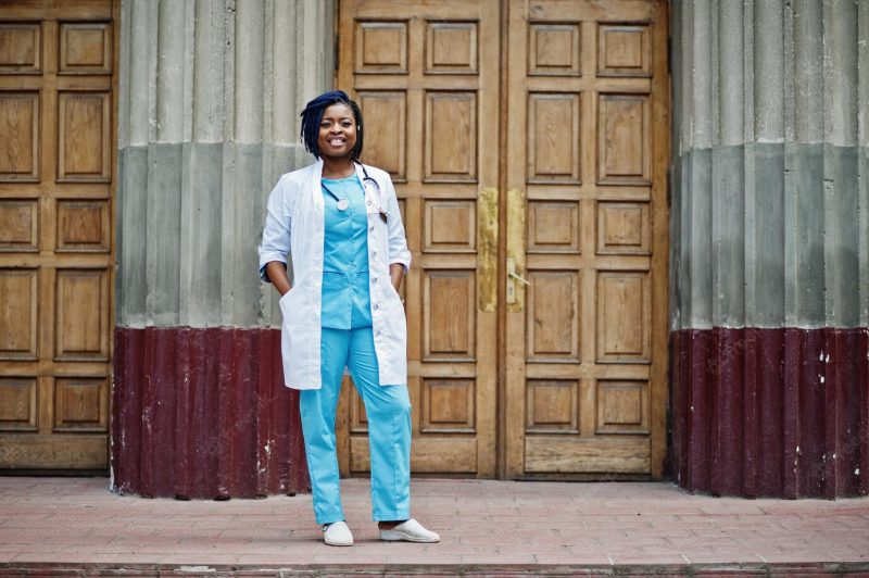 Stylish african american doctor with stethoscope and lab coat posed against door of hospital Free Photo