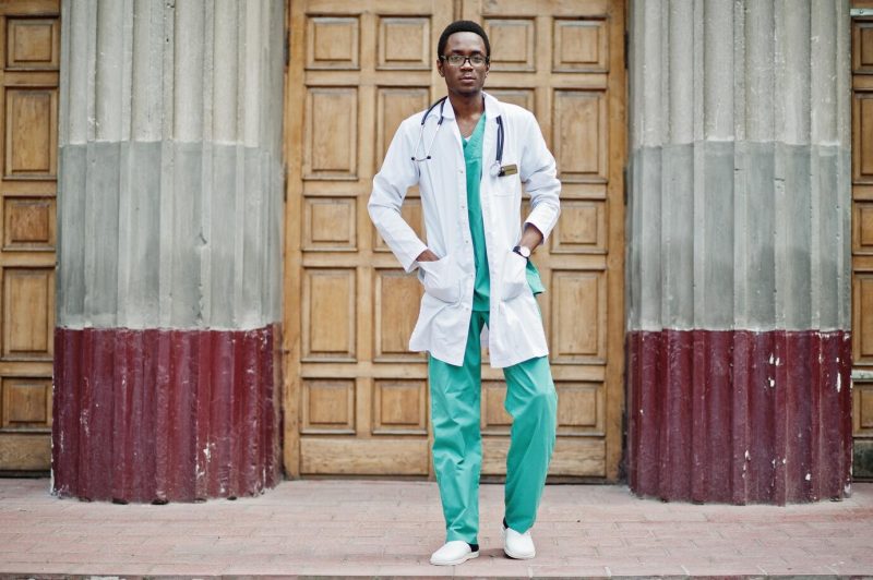 Stylish african american doctor with stethoscope and lab coat posed against door of hospital Free Photo