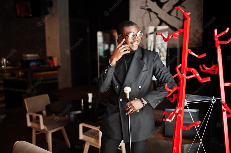 Stylish african american gentleman in elegant black jacket and glasses holding retro walking stick as cane flask or tippling cane with golden diamond ball handle and speaking on mobile phone Free Photo