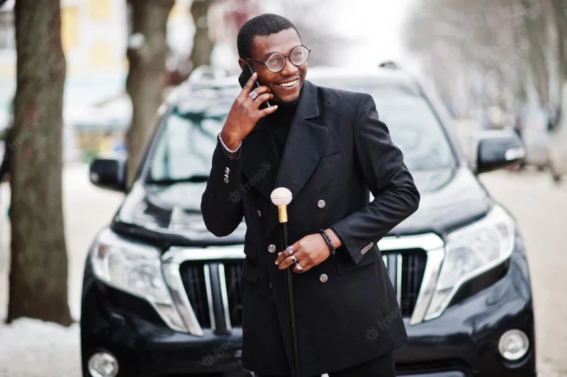 Stylish African American gentleman in elegant black jacket holding retro walking stick as cane flask or tippling cane rich fashionable afro man against business suv car speaking on mobile phone Free Photo