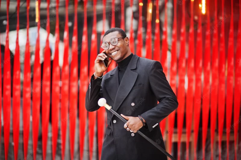 Stylish african american gentleman in elegant black jacket holding retro walking stick as cane flask or tippling cane rich fashionable afro man against red wall speaking on mobile phone Free Photo