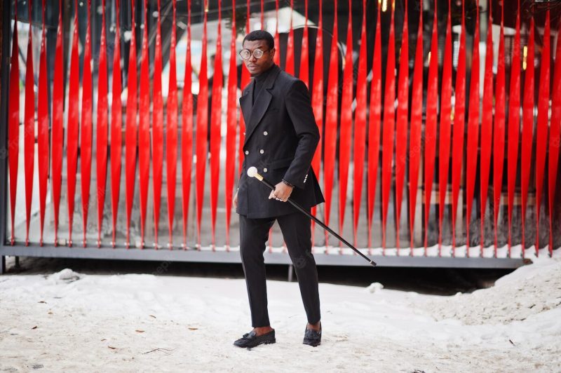 Stylish african american gentleman in elegant black jacket holding retro walking stick as cane flask or tippling cane with golden diamond ball handle rich fashionable Afro man against red wall Free Photo