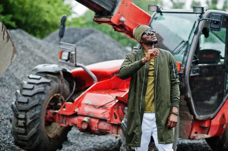 Stylish African American man in hat and sunglasses posed outdoor in rain against tractor with a bucket Free Photo