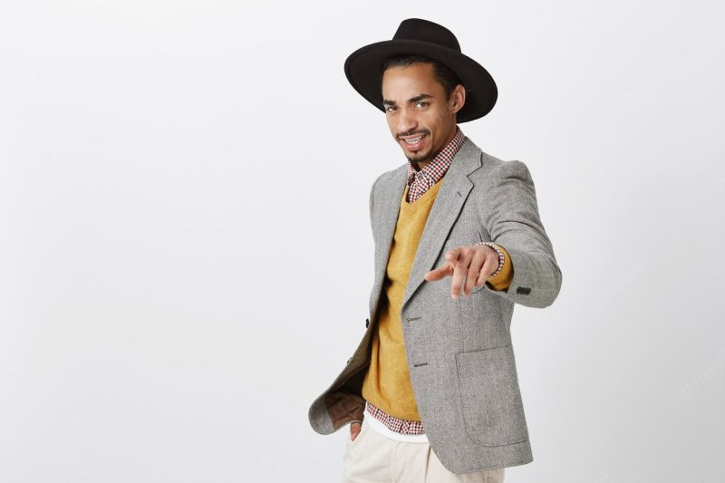 Stylish employee talking with employers, giving new tasks. confident fashionable dark-skinned male model in trendy black hat and gray jacket pointing, being self-assured over gray wall Free Photo