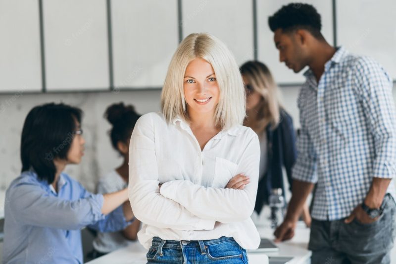 Stylish female secretary standing in confident pose in conference hall and smiling. indoor portrait of pretty blonde office worker waiting for negotiation with partners. Free Photo