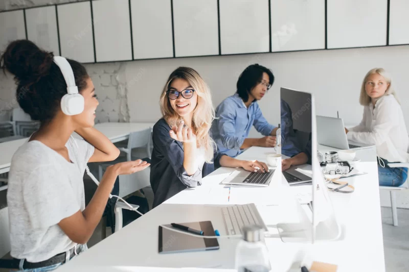 Stylish female web-programmers talking about work while spending time in office. indoor portrait of african woman in headphones and Asian worker using computers. Free Photo