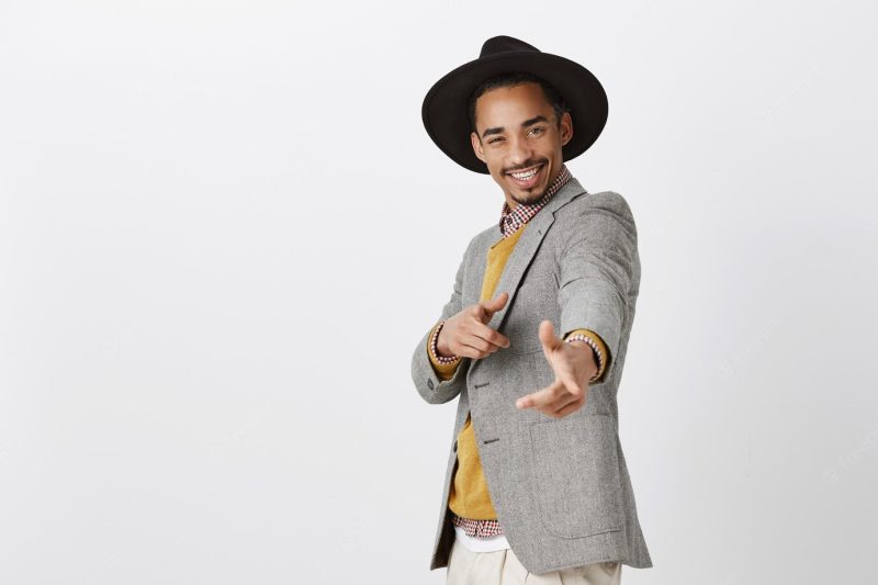 Stylish man charmed girls with his charisma. indoor shot of good-looking stylish male in formal clothes and hat, pointing and smiling broadly, being in happy and good mood over gray wall Free Photo