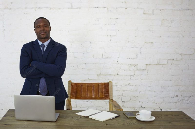 Stylish successful african american male entrepreneur having confident look standing at his workplace against white brick wall with copy space for your text or promotional information Free Photo