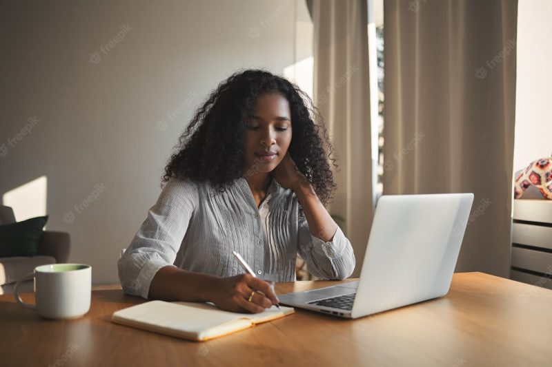 Successful attractive young Afro American businesswoman in stylish shirt sitting at her workplace in front of open portable computer and making notes in her diary, having thoughtful facial expression Free Photo