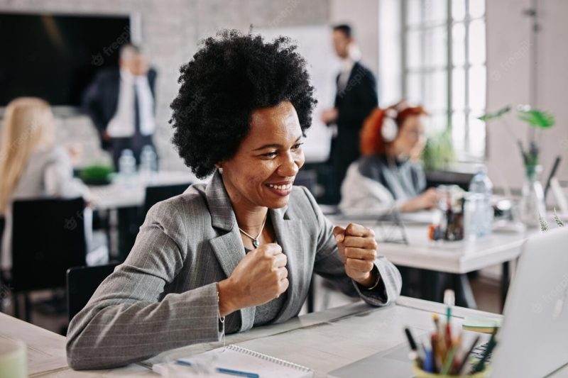 Successful black businesswoman working on a computer and celebrating in the office there are people in the background Free Photo