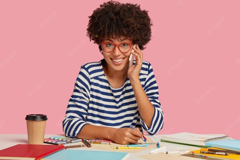 Successful dark skinned woman with afro haircut, dressed in striped clothes, has pleasant telephone conversation while draws something in notepad, drink coffee to go, feels satisfied and inspired Free Photo