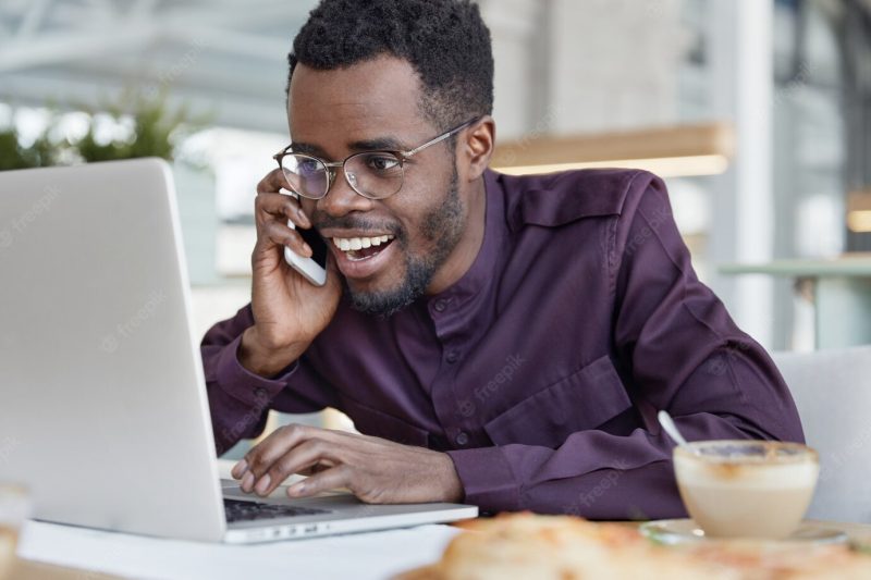 Successful happy dark skinned african male executive, smiles happily and looks at laptop computer Free Photo