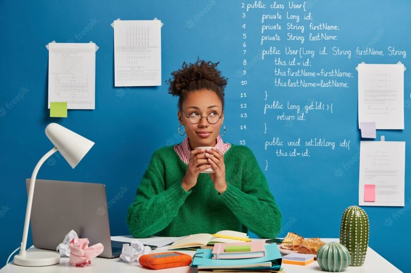 Successful woman copywriter works on online project, looks thoughtfully aside, drinks aromatic coffee, poses in cozy study room Free Photo