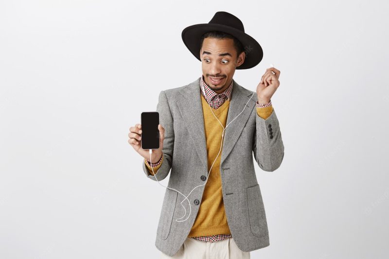 Surprised African-American guy take-off headphone ambushed and awkward, looking at smartphone screen, showing mobile display Free Photo