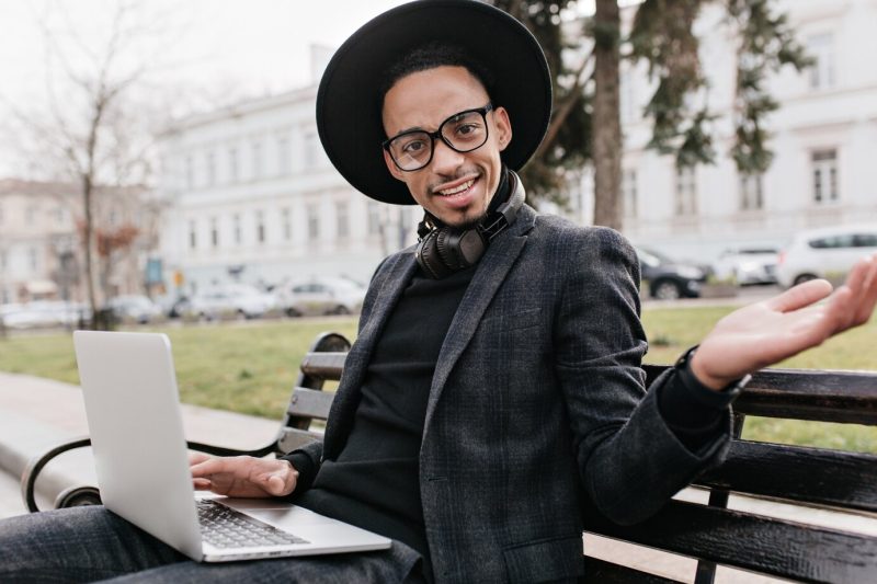 Surprised african student sitting on bench with computer. outdoor photo of funny black male freelancer using laptop for work. Free Photo