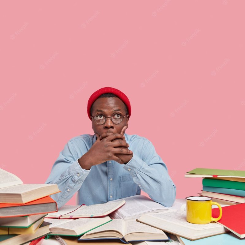 Surprised dark skinned man covers mouth with both hands, tries be speechless, focused upwards, wears red hat and shirt, poses at desktop Free Photo