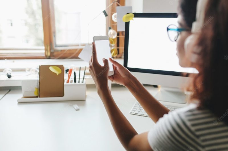 Tanned girl in striped summer shirt using smartphone sitting at the table with computer on it Free Photo