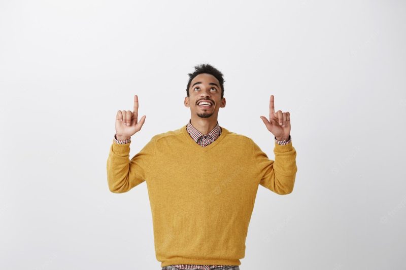 Thanks god it is friday. portrait of pleased interested young African-American student in stylish yellow sweater raising hands, looking and pointing up, enjoying nice view of sky Free Photo